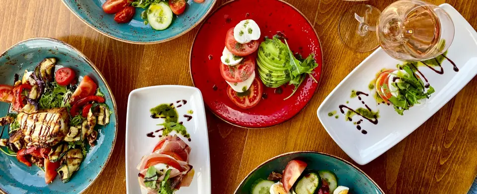 Plates of burrata, salads and cured meats on turquoise and red glazed plates, seen from above on a wooden table